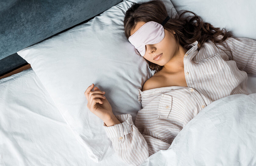 Ung brunett kvinna med sovmask vilar i sängen i ljus pyjamas