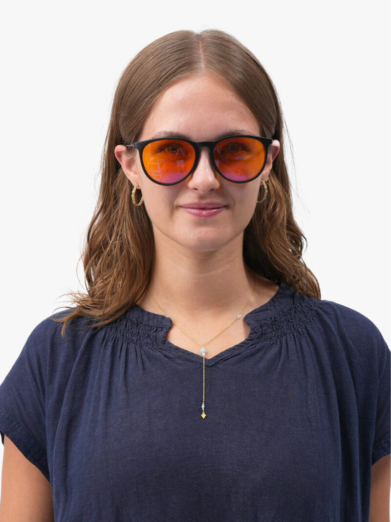 Kvinna med svarta bågar och orange spegelglasögon, guldfärgade hoops, delikat lariat-halsband och marinblå blus