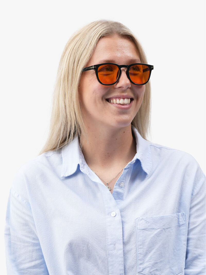 Leende kvinna med blont hår i ljusblå skjorta bär svarta solglasögon med orangea glas