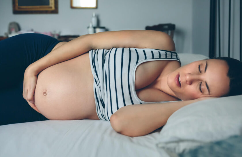 Gravid kvinna sover på sidan i sängen med handen på magen, bär randigt linne
