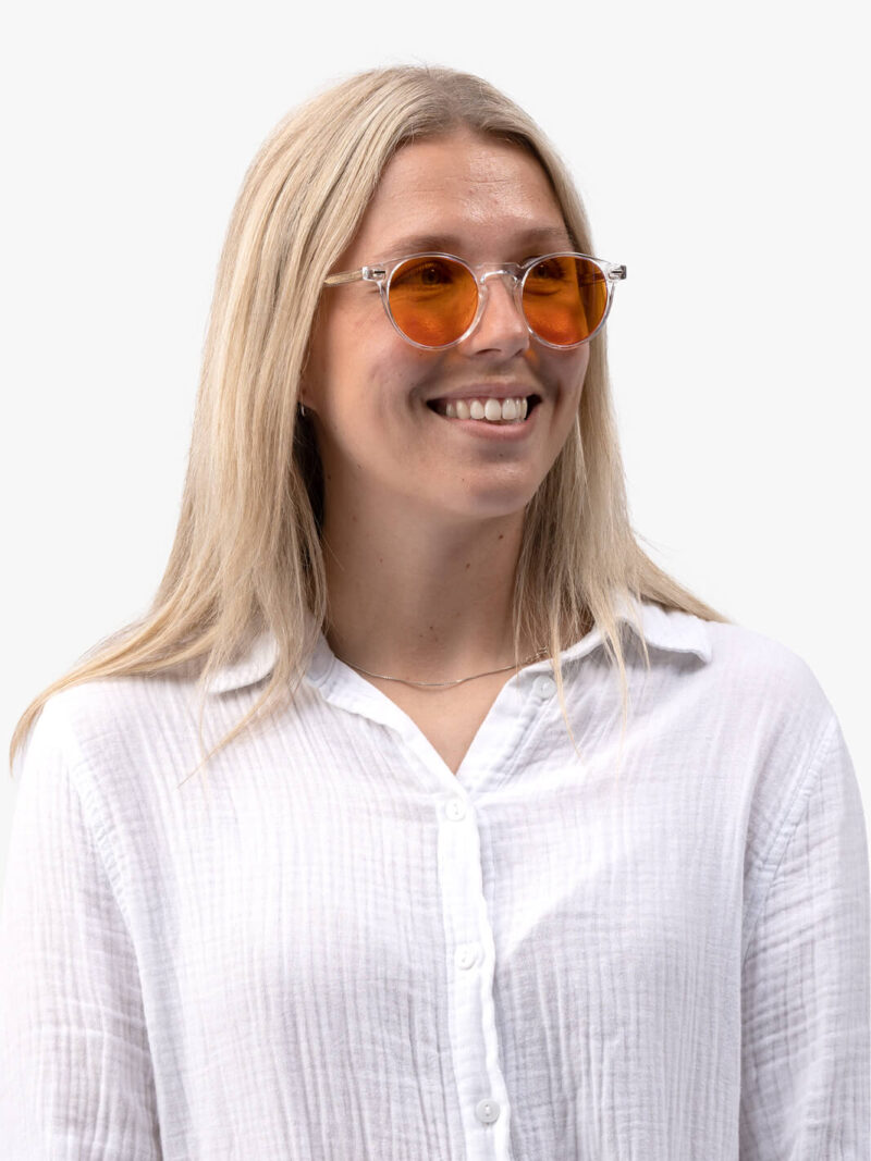 Leende ung kvinna med långt blont hår, runda orange solglasögon och vit skjorta, vänd åt sidan