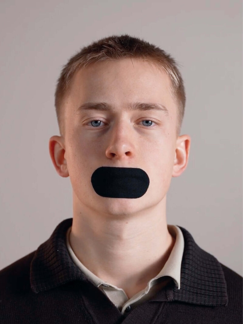 Ung man med kort blont hår och svart muntejp över munnen mot neutral bakgrund