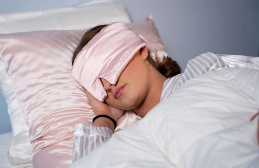 Kvinna sover med rosa silkessovmask och matchande satinörngott på vit bäddning