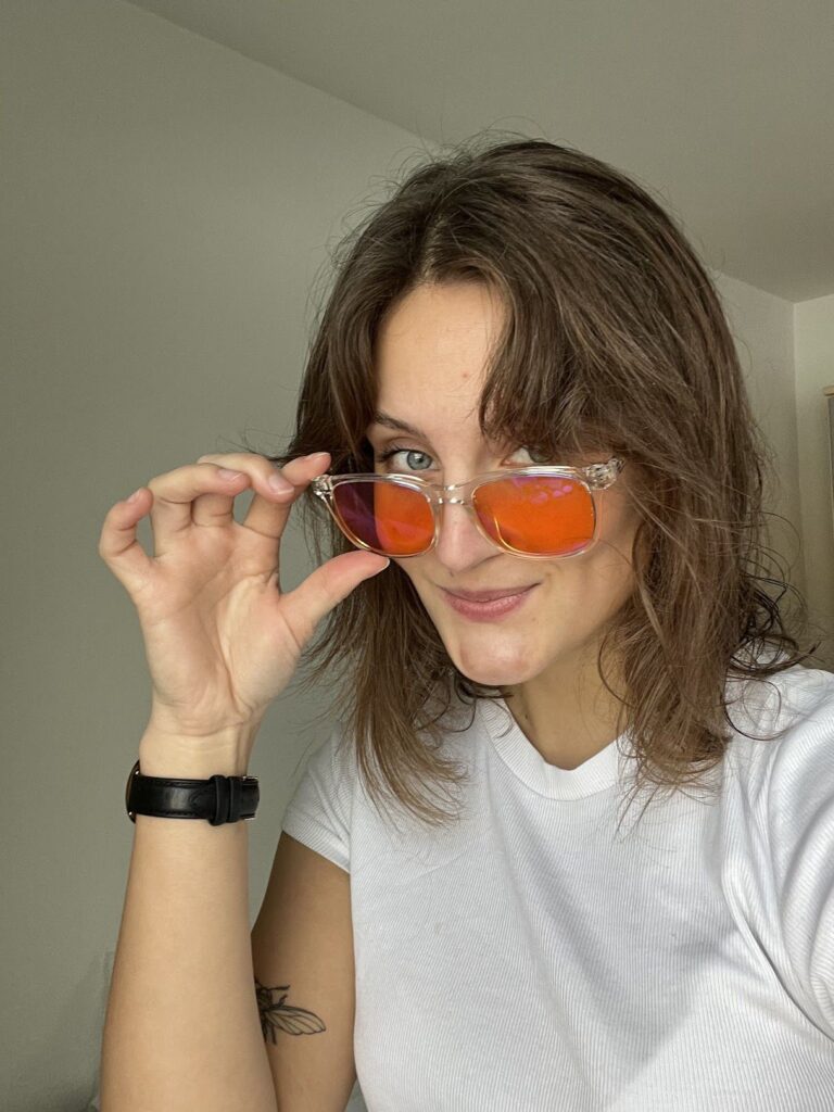 Leende kvinna i vit t-shirt som håller upp solglasögon med orangea spegelglas, synlig klocka och insektstatuering på armen