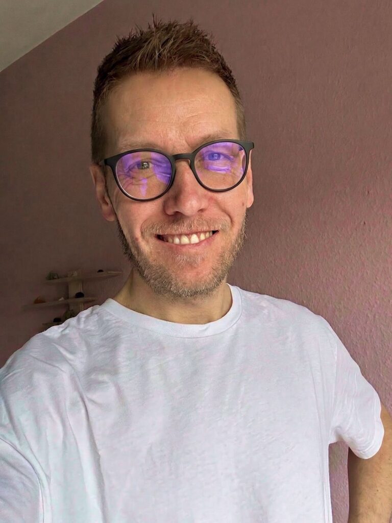 Leende man i vit t-shirt med runda glasögon som reflekterar blått ljus framför rosa vägg